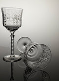A clear cut-glass wine glass with an etched floral pattern and a ribbed stem, shown standing upright alongside a matching glass viewed from the side on a reflective gray surface.