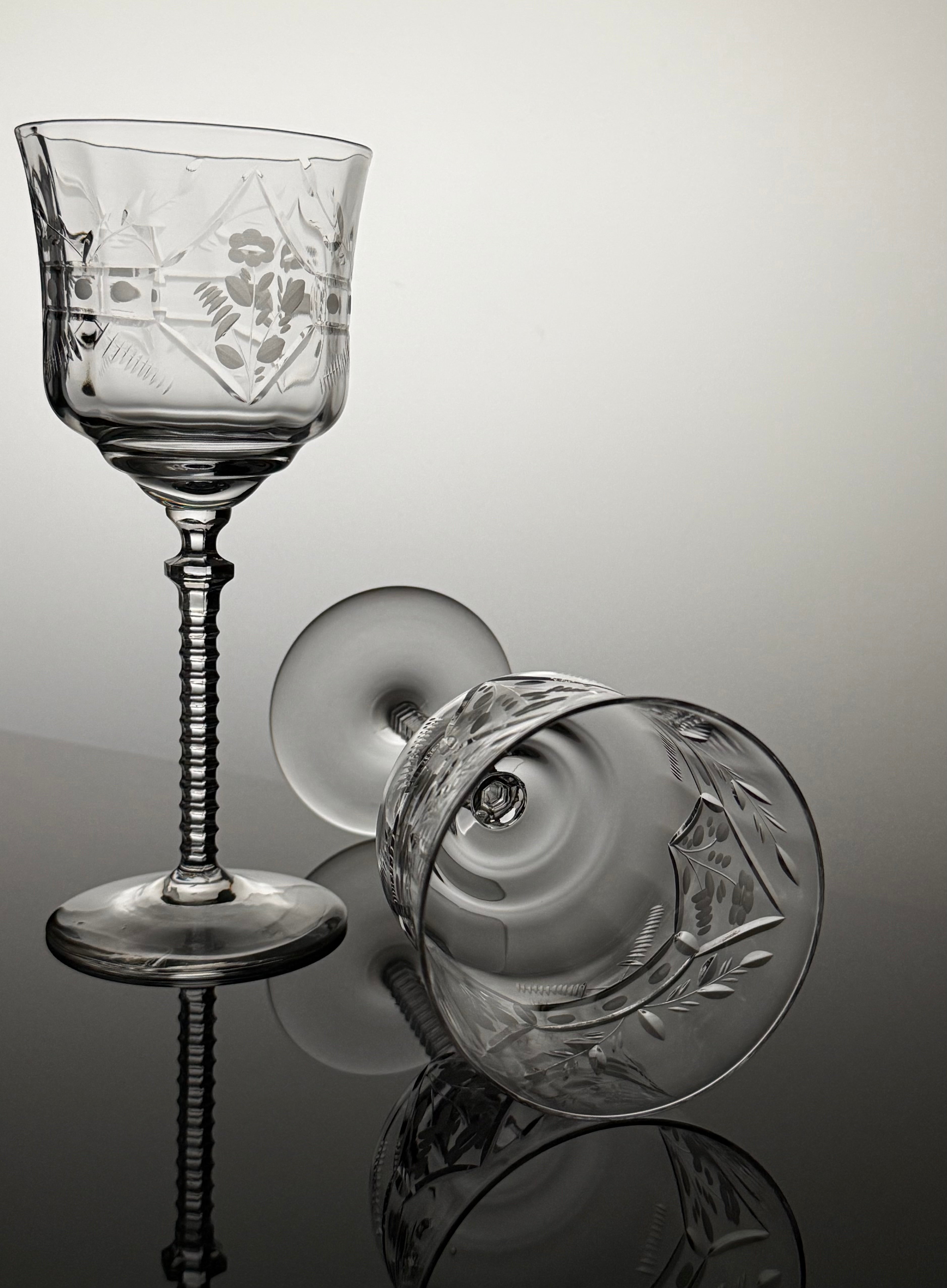 A clear cut-glass wine glass with an etched floral pattern and a ribbed stem, shown standing upright alongside a matching glass viewed from the side on a reflective gray surface.