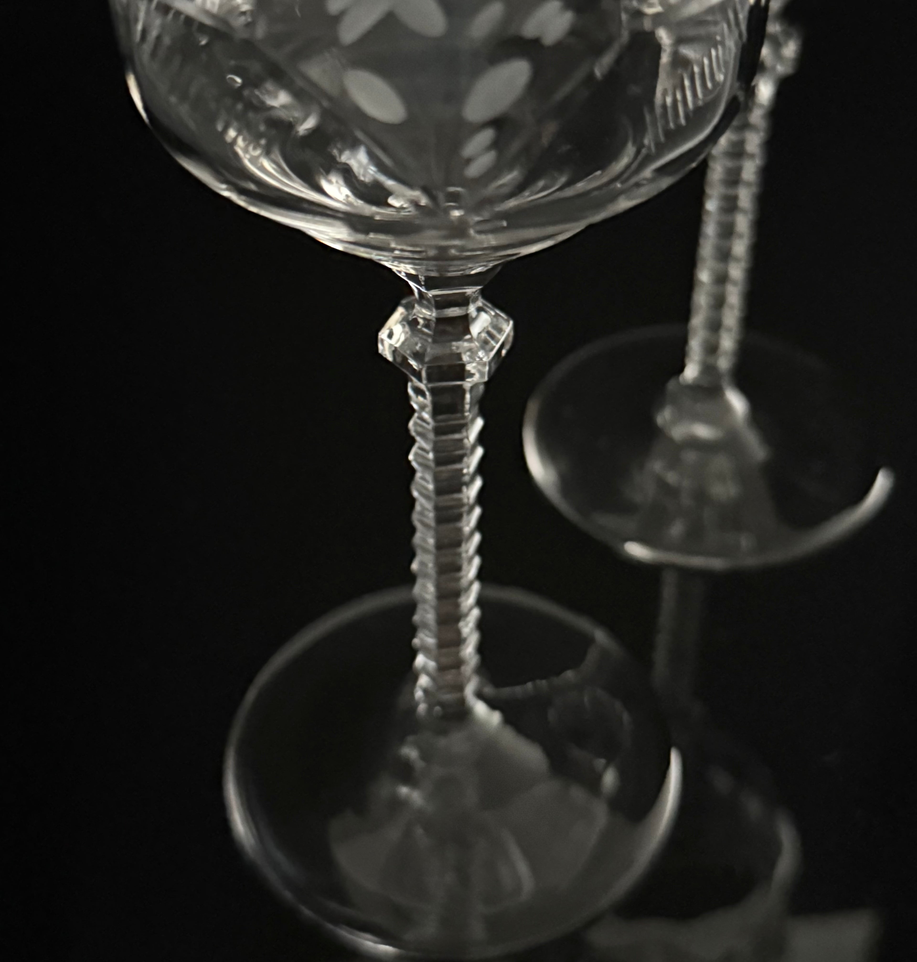 A close-up of a clear cut-glass or crystal stemmed glass with a flared base and decorative etched pattern, photographed against a black background with another similar glass blurred in the background.