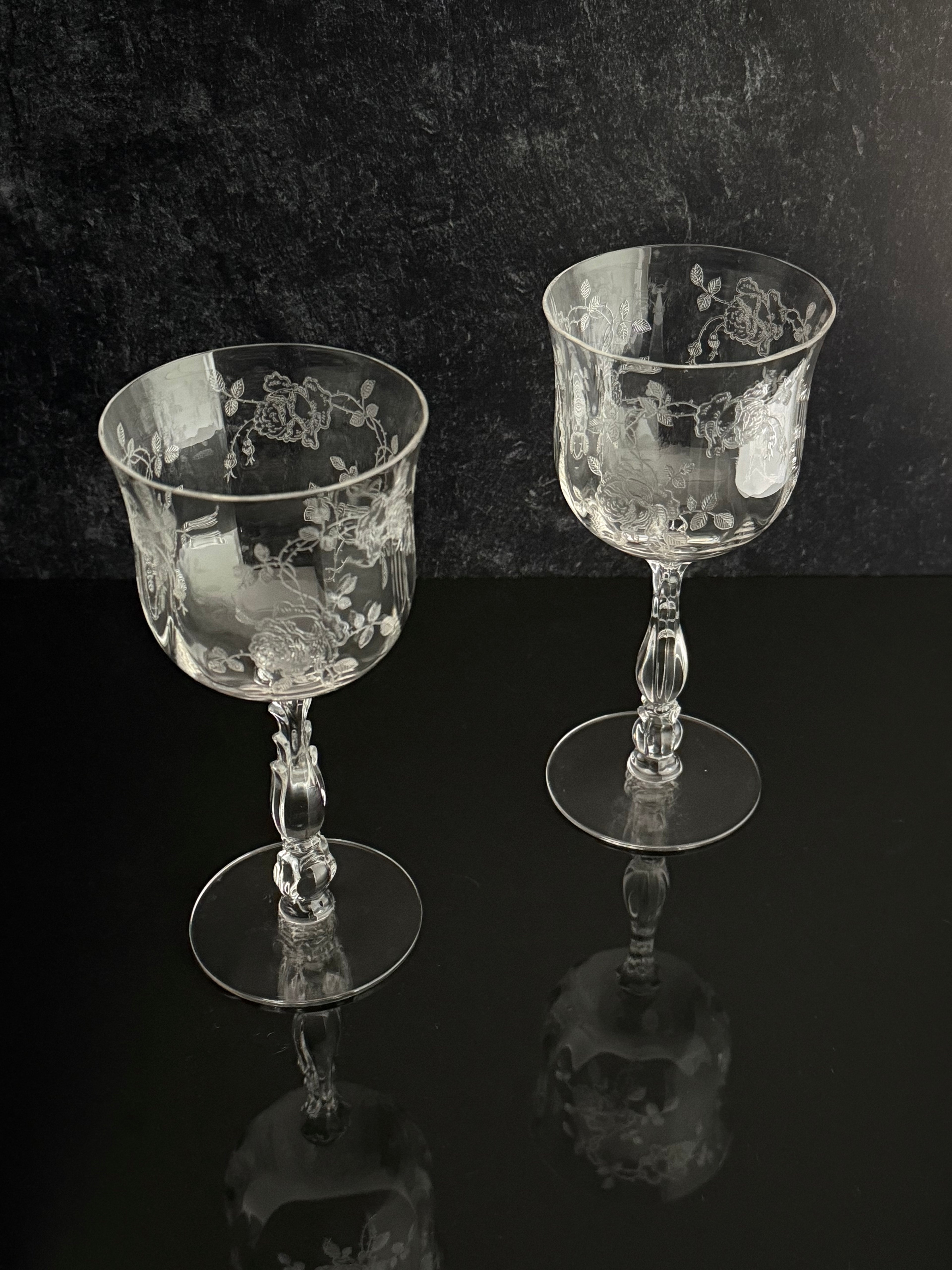 Two clear glass goblets with etched floral patterns and flared circular bases, displayed on a dark reflective surface against a dark textured background.