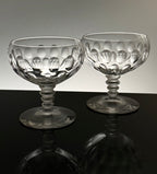 Two clear cut-glass coupe glasses with a textured, patterned bowl and flared stems, displayed on a reflective black surface against a light gray background.