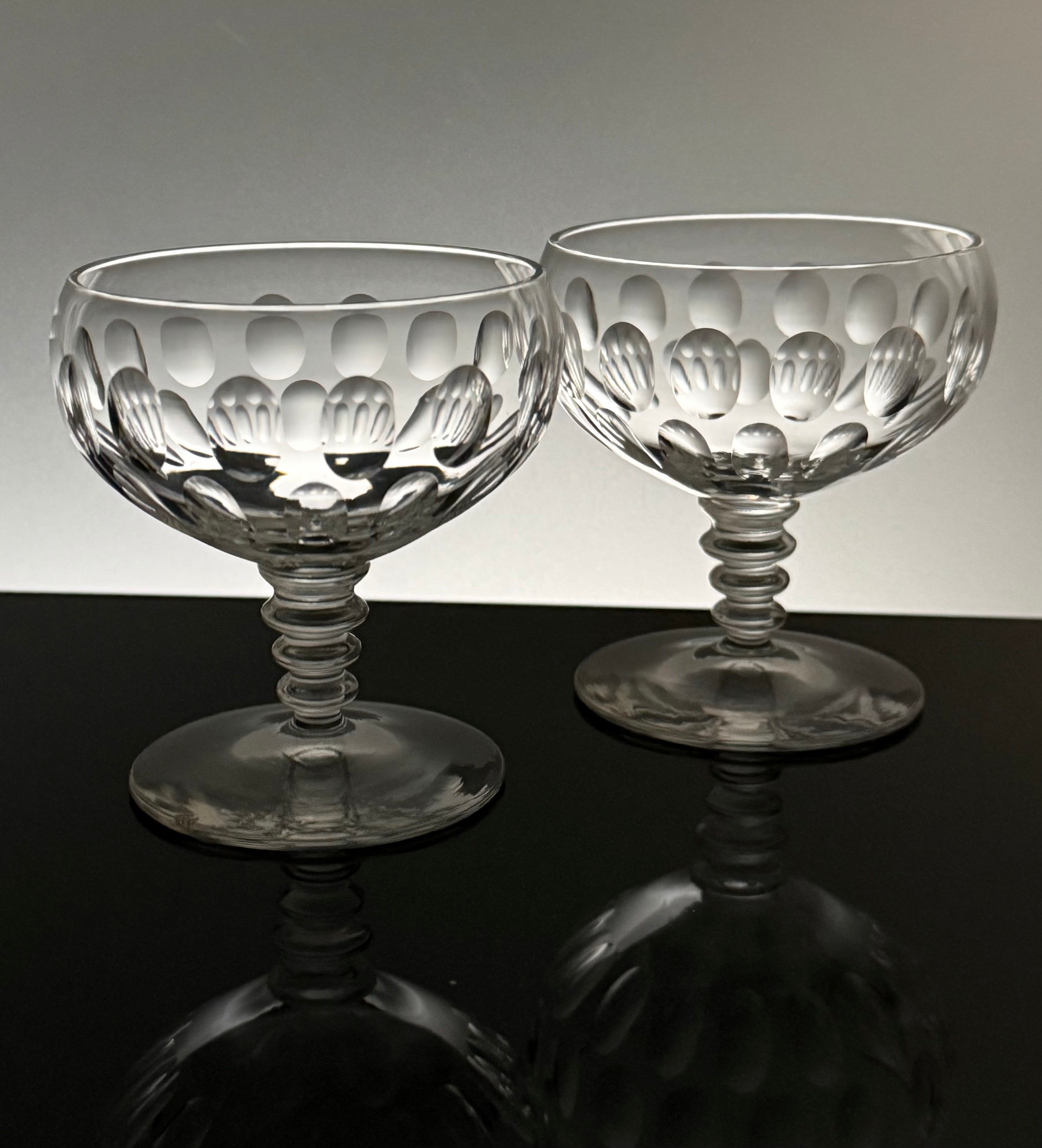 Two clear cut-glass coupe glasses with a textured, patterned bowl and flared stems, displayed on a reflective black surface against a light gray background.