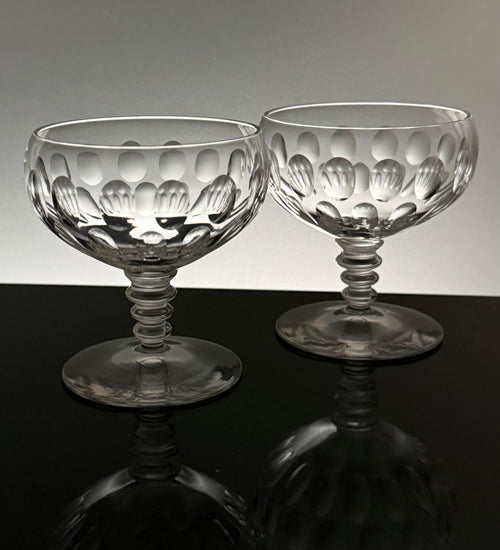 Two clear cut-glass coupe glasses with a textured, patterned bowl and flared stems, displayed on a reflective black surface against a light gray background.