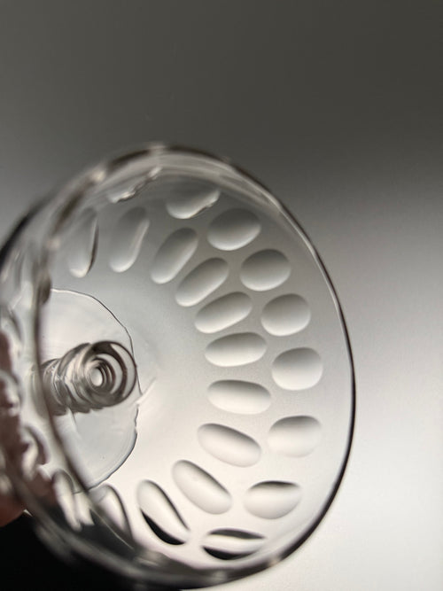 A close-up of a clear glass coupe glass with a textured, perforated oval pattern on the bowl, held by a hand against a dark gray gradient background.