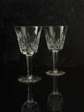 Two clear cut-glass wine glasses with stems and circular bases, standing on a dark reflective surface against a dark textured background.