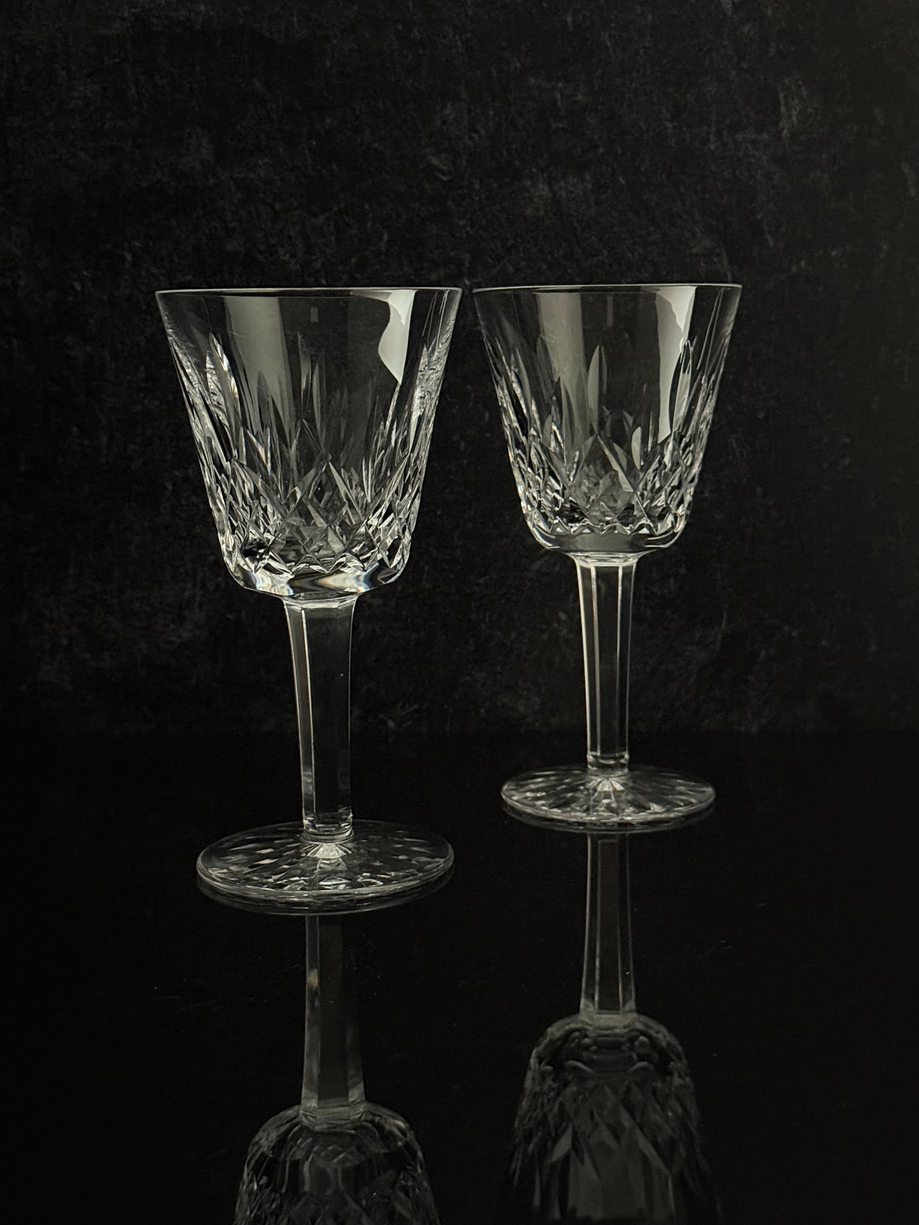 Two clear cut-glass wine glasses with stems and circular bases, standing on a dark reflective surface against a dark textured background.