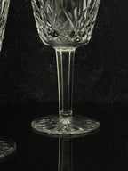A close-up of a clear cut-crystal wine glass with a faceted stem and circular base, set against a dark reflective background.