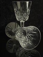 A clear cut-crystal wine glass and a matching coupe-style glass with faceted starburst patterns, arranged on a dark reflective surface against a black background.
