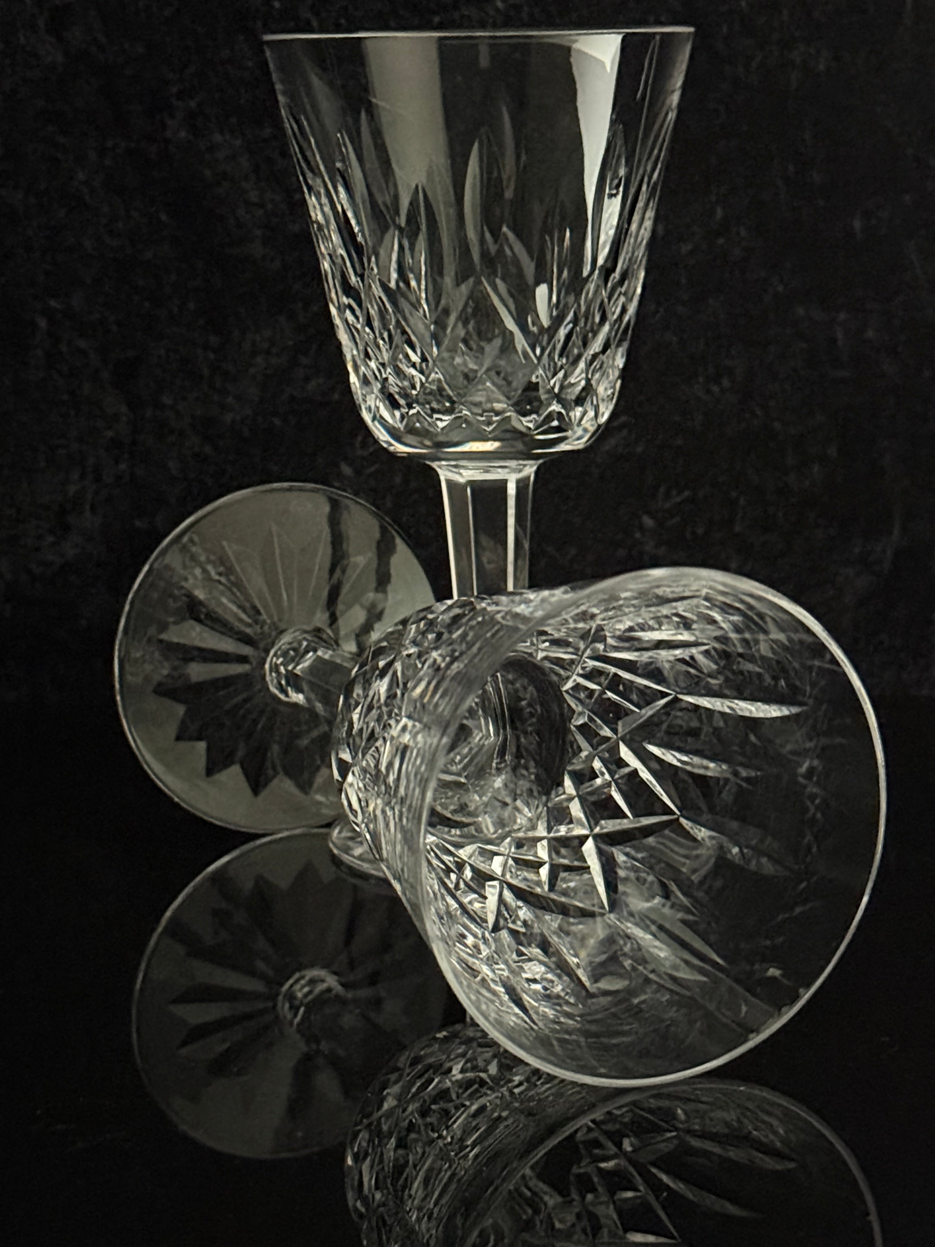 A clear cut-crystal wine glass and a matching coupe-style glass with faceted starburst patterns, arranged on a dark reflective surface against a black background.