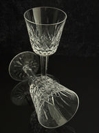 A clear cut-crystal wine glass with a faceted stem, shown upright with its reflection visible on a glossy black surface against a dark background.