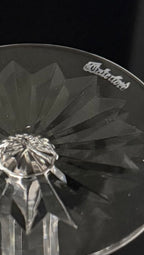 A close-up of a clear cut-glass or crystal glass with a faceted geometric pattern and a visible 'Waterford' logo near the rim, photographed against a dark background.