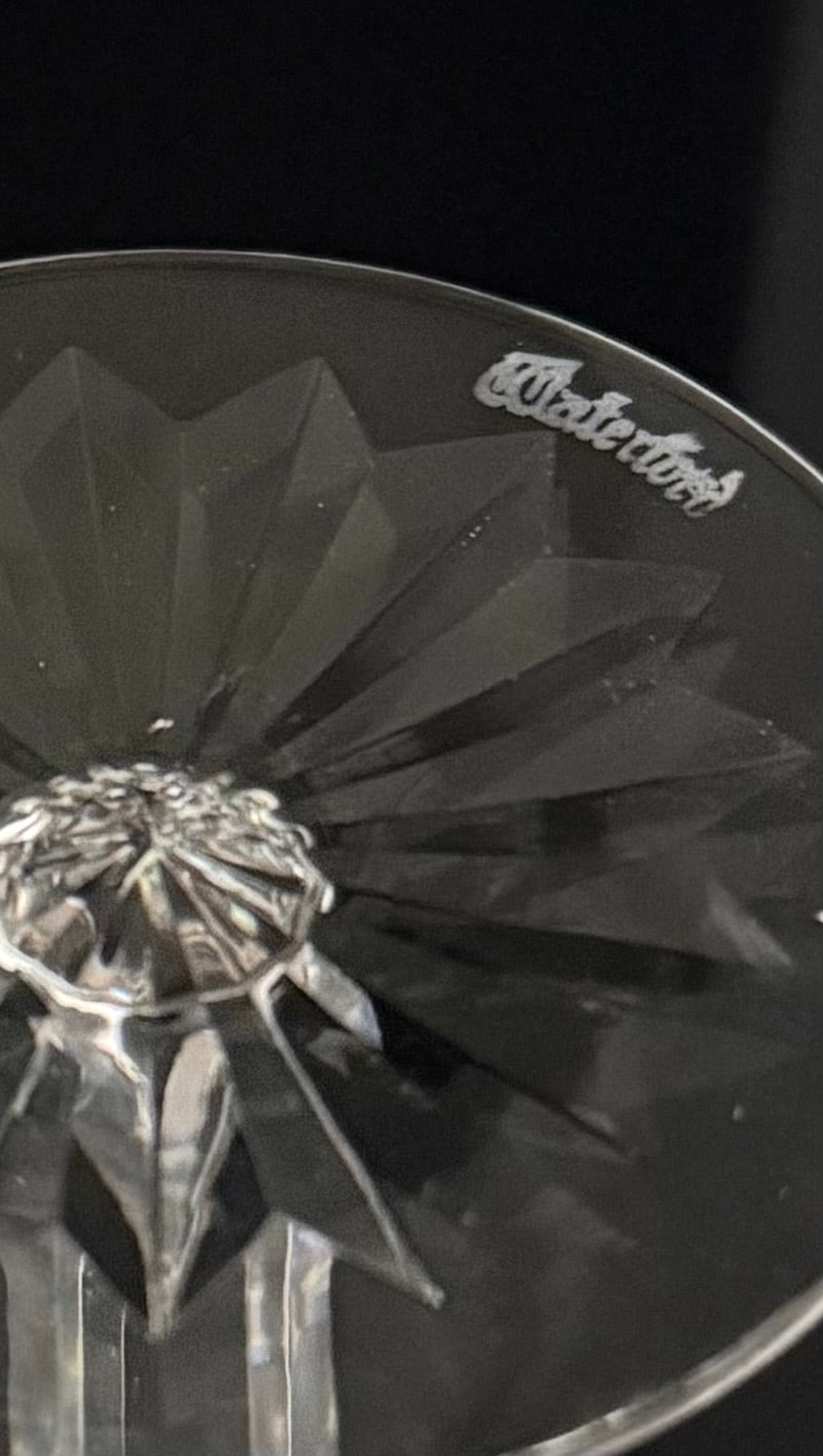 A close-up of a clear cut-glass or crystal glass with a faceted geometric pattern and a visible 'Waterford' logo near the rim, photographed against a dark background.