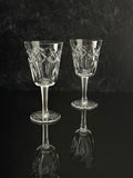 Two clear cut-glass wine glasses with geometric patterns on the bowls and stems, standing on a dark reflective surface against a dark textured background.