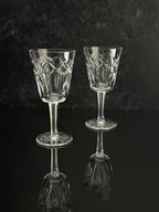 Two clear cut-glass wine glasses with geometric patterns on the bowls and stems, standing on a dark reflective surface against a dark textured background.