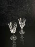 Two clear cut-crystal wine glasses with faceted stems and round bases, standing on a dark reflective surface against a dark textured background.
