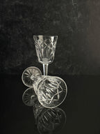 A clear cut-crystal wine glass standing upright, with a second matching glass lying on its side in front, set against a dark reflective background.