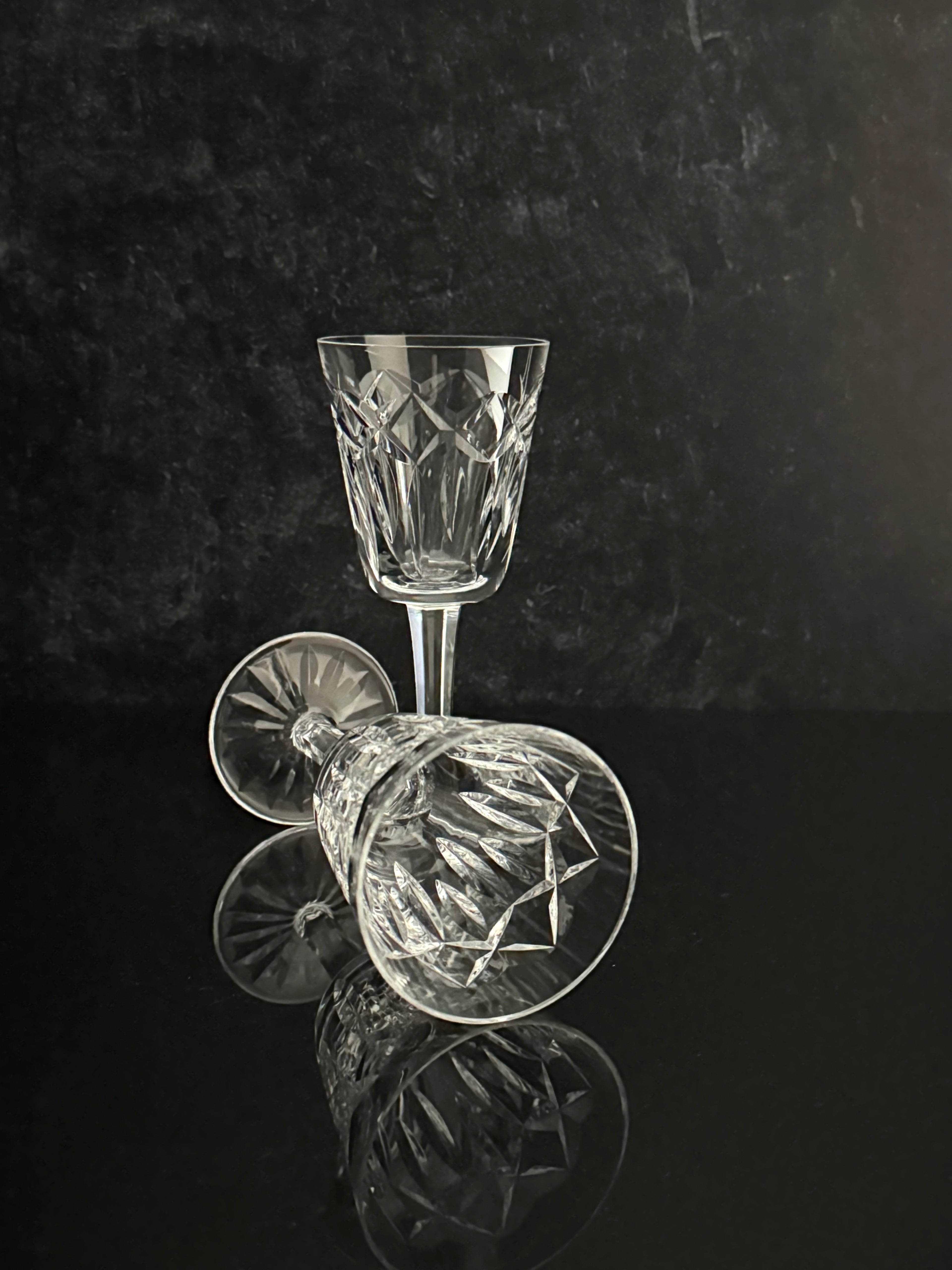 A clear cut-crystal wine glass standing upright, with a second matching glass lying on its side in front, set against a dark reflective background.