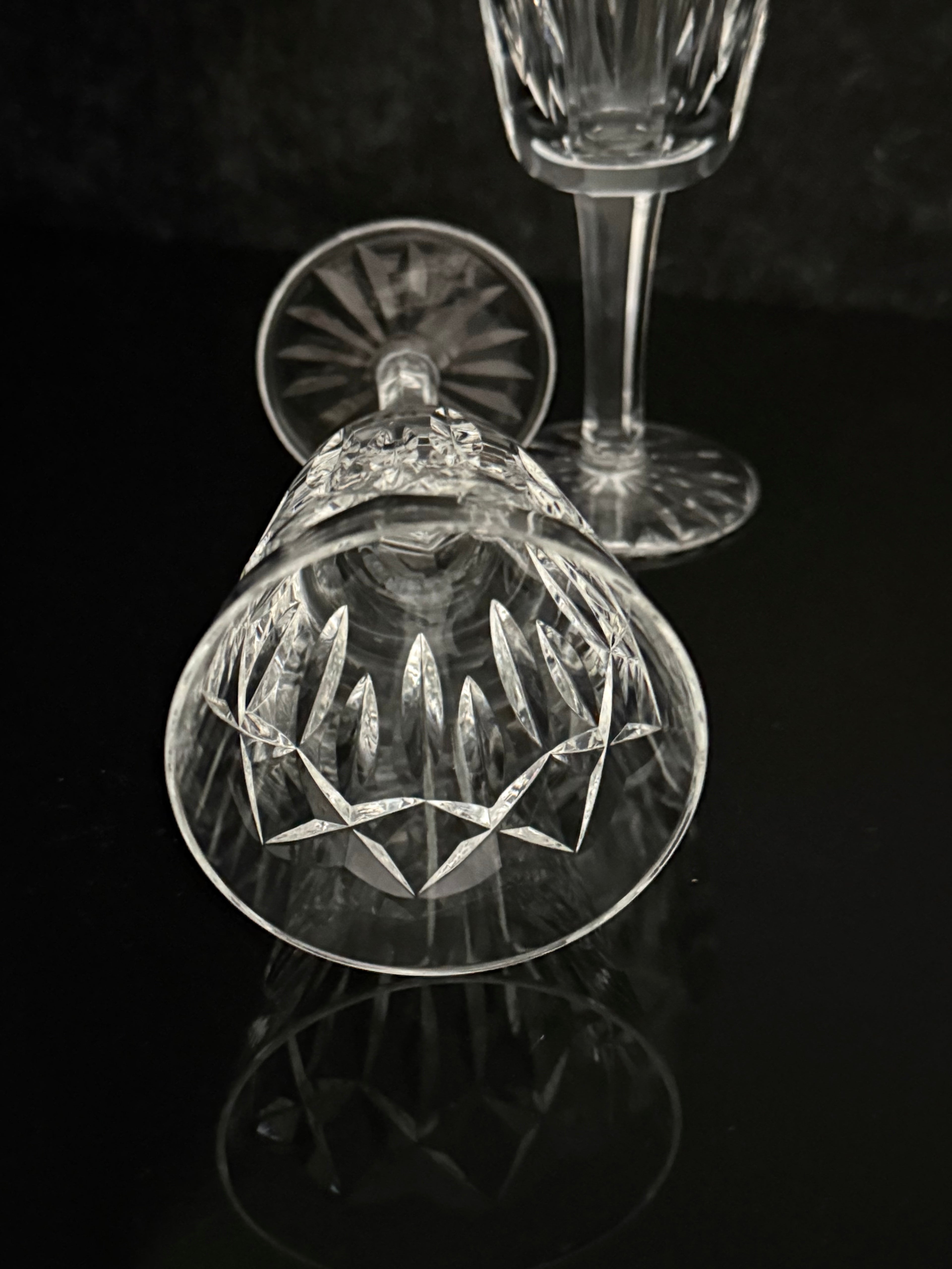 A clear cut-crystal wine glass shown upside down on a glossy black reflective surface, with another crystal glass standing behind it against a dark background.