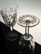 Two clear cut-crystal wine glasses with geometric and floral starburst patterns, one standing upright and the other tilted to show the bowl and stem, on a black and white background.
