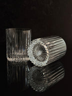 Two clear cut-crystal tumblers with vertical and radial faceted patterns, one standing upright and the other lying on its side, set against a dark reflective background.