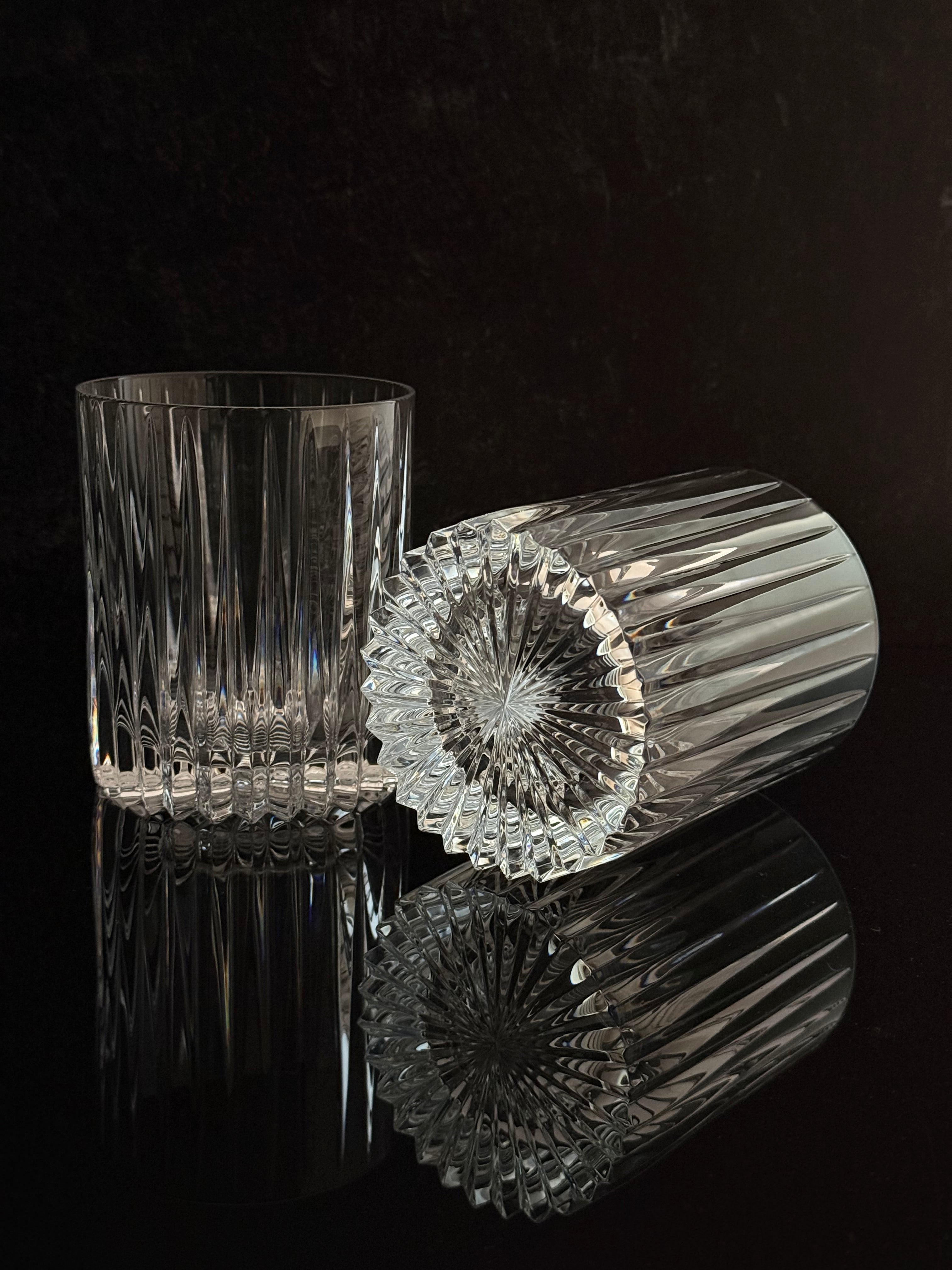 Two clear cut-crystal tumblers with vertical and radial faceted patterns, one standing upright and the other lying on its side, set against a dark reflective background.