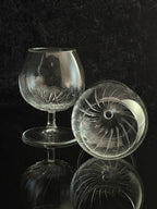 A clear cut-glass or crystal snifter glass with a stem and round base, shown alongside a matching clear glass dome lid with radial cut patterns, set against a dark reflective background.