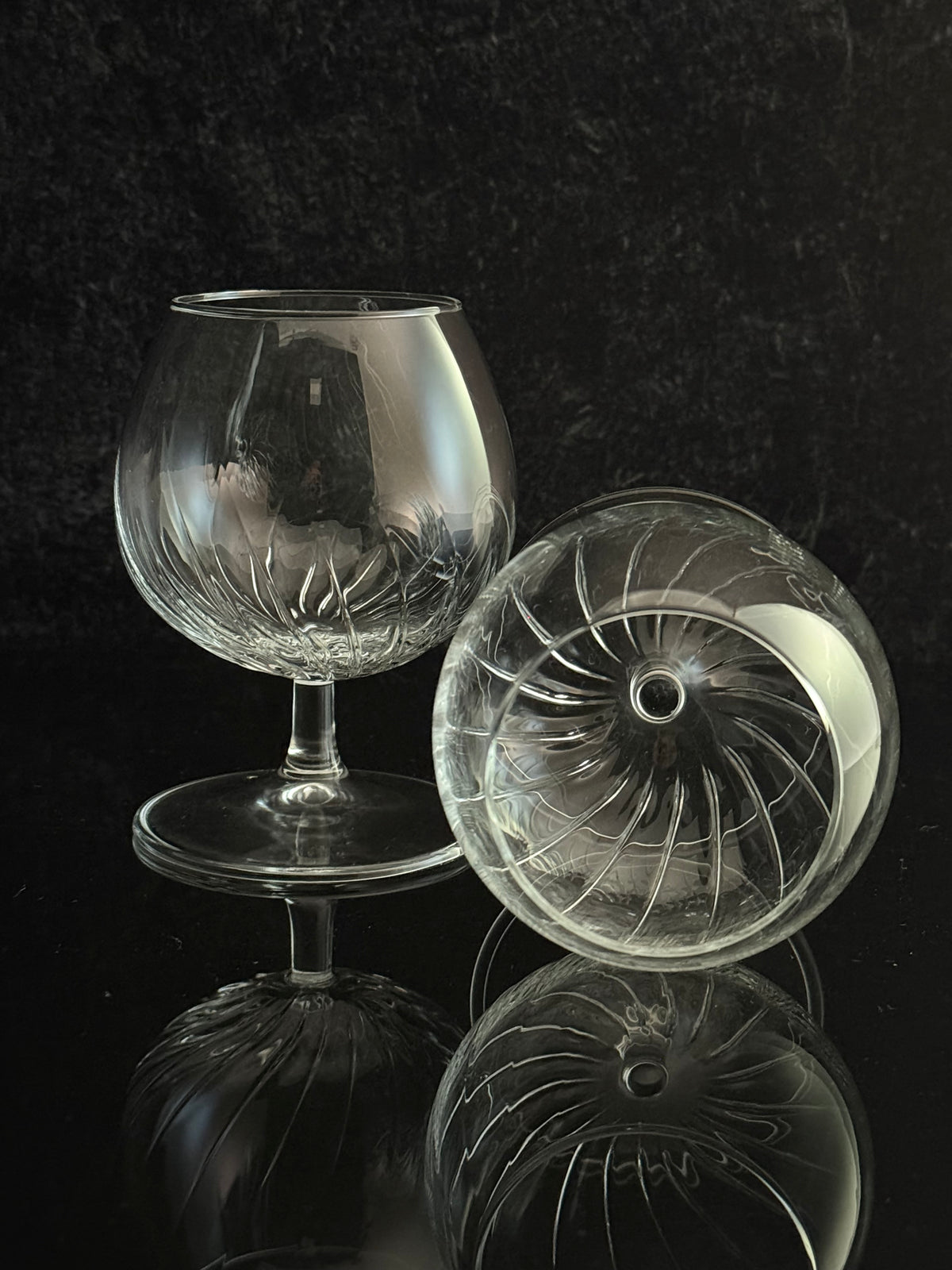 A clear cut-glass or crystal snifter glass with a stem and round base, shown alongside a matching clear glass dome lid with radial cut patterns, set against a dark reflective background.