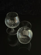 Two clear cut-glass or crystal snifter glasses with stem bases, displayed on a glossy black reflective surface against a dark background.