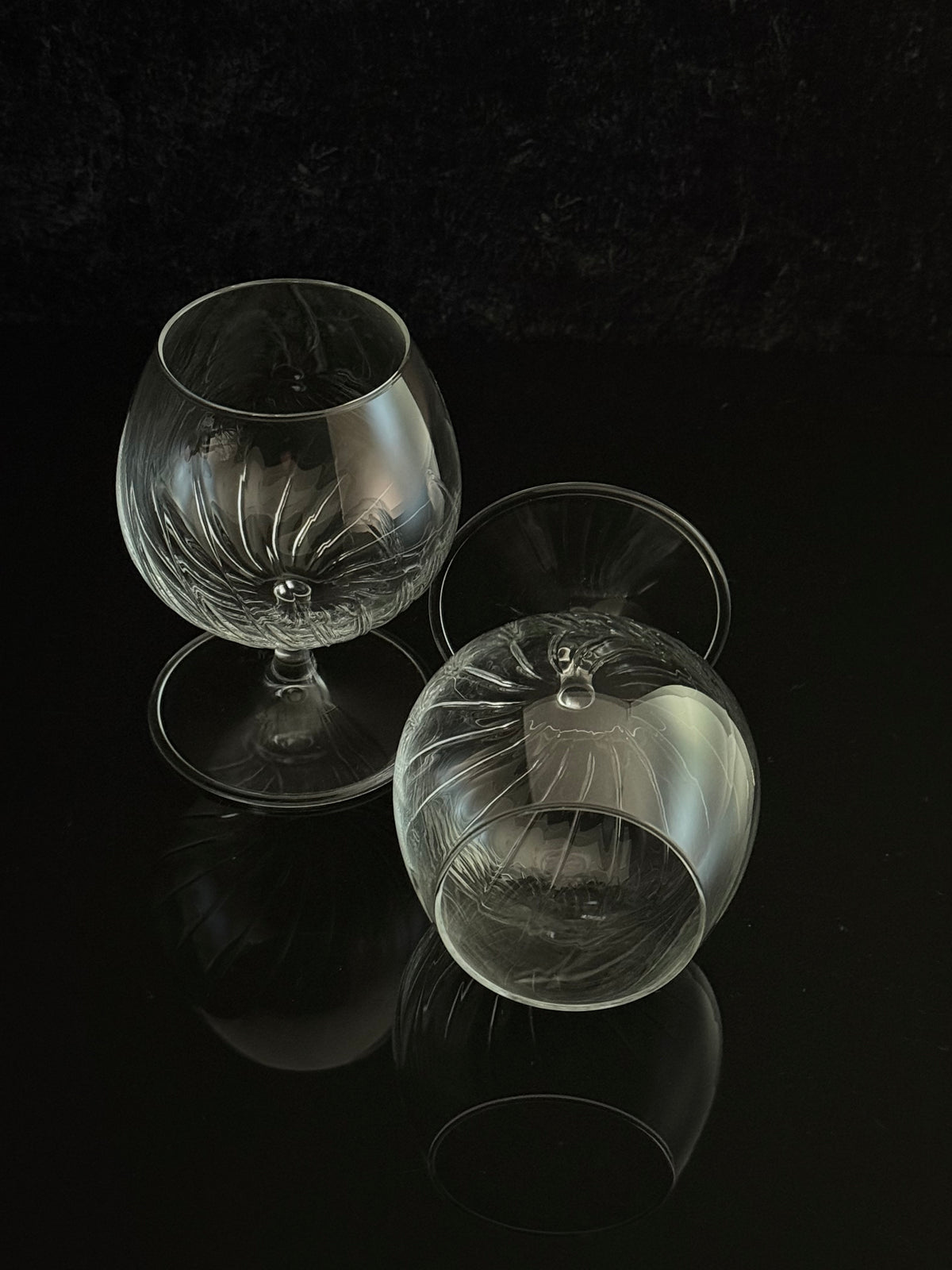 Two clear cut-glass or crystal snifter glasses with stem bases, displayed on a glossy black reflective surface against a dark background.