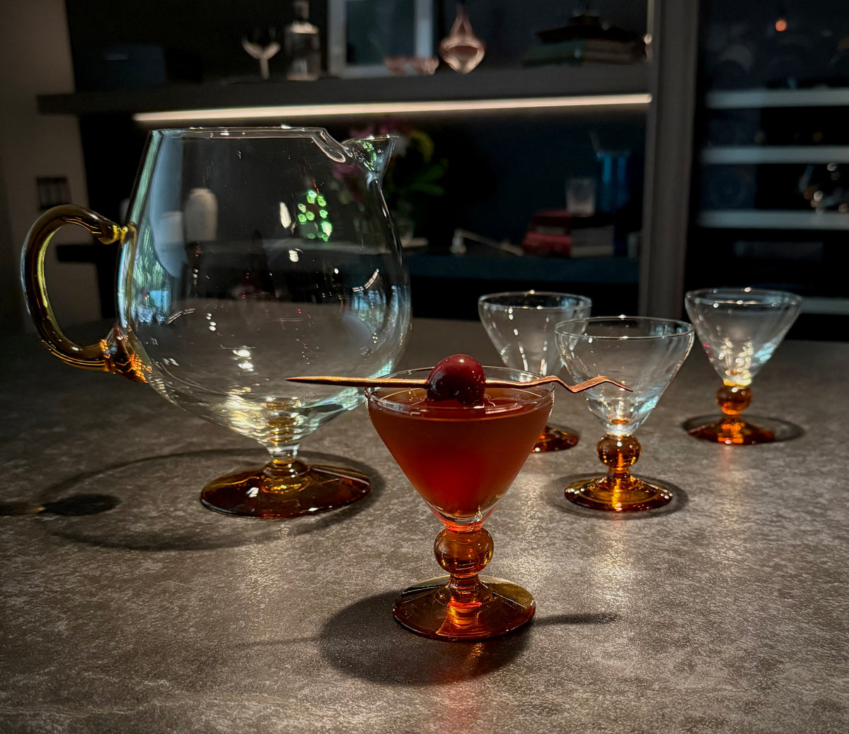 A clear glass cocktail pitcher with amber-colored base and handle, shown with several matching coupe glasses on a dark countertop in a dimly lit bar or kitchen setting.