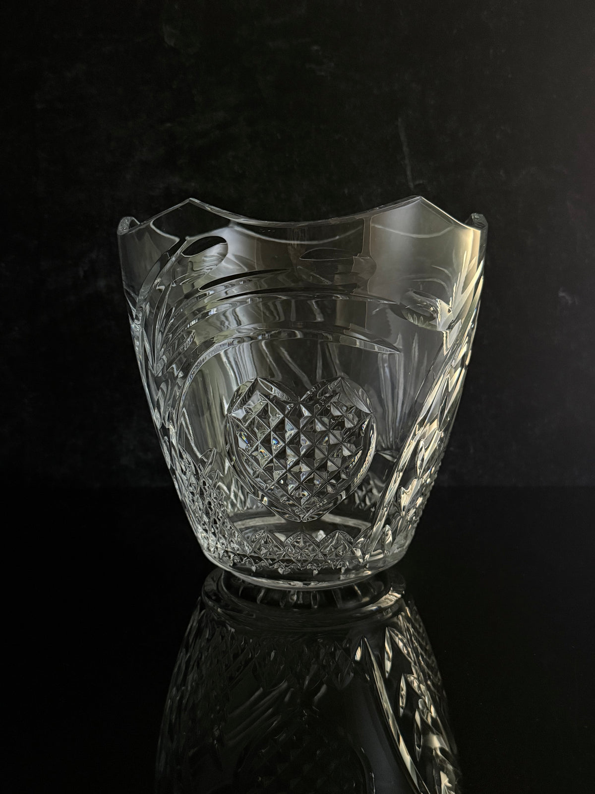 A clear cut-crystal ice bucket with a diamond-patterned center and wavy rim, shown on a reflective black surface against a dark background.