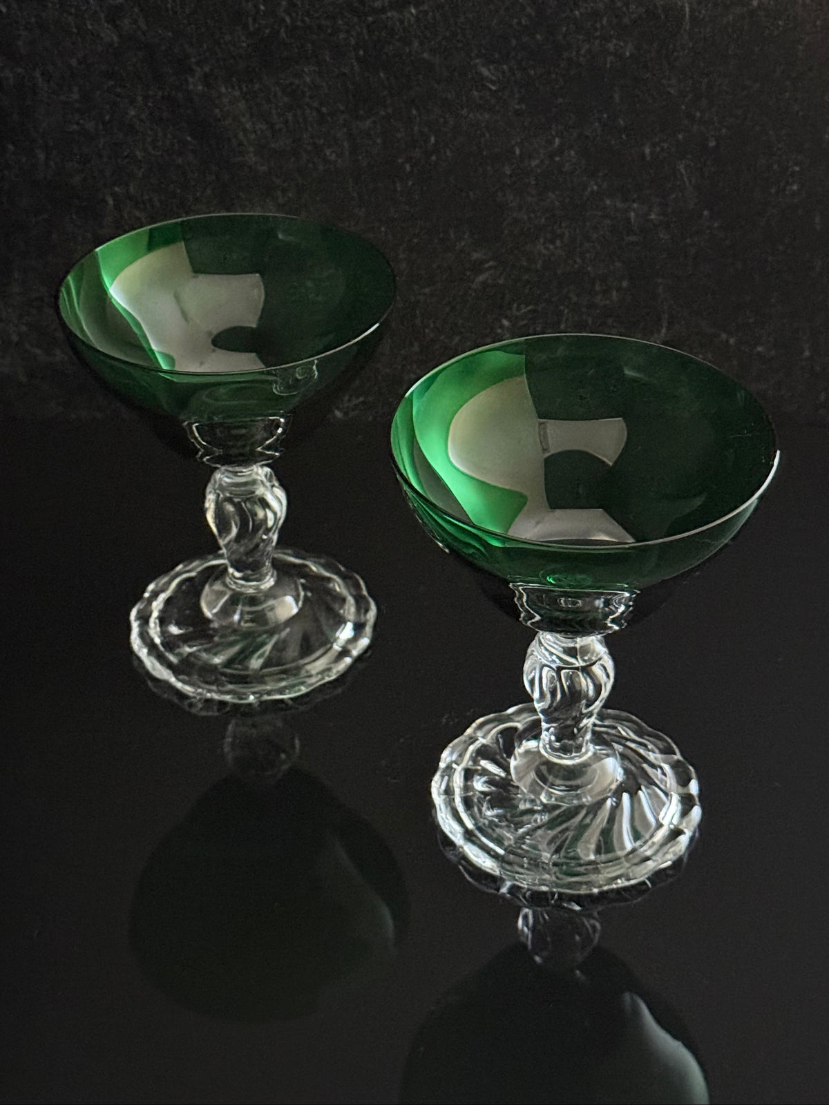 Two elegant coupe glasses with deep green bowls and clear glass stems and bases, displayed on a reflective black surface against a dark textured background.