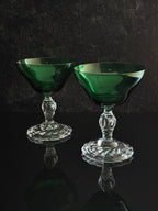 Two green glass coupe cups with clear cut crystal stems and bases, displayed on a reflective black surface against a dark textured background.