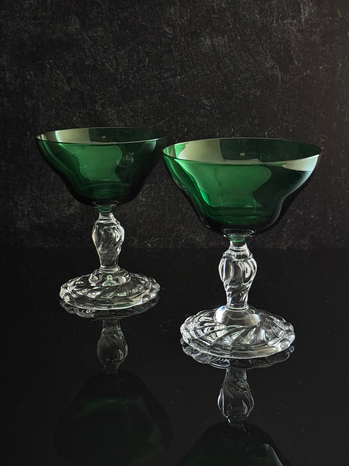 Two green glass coupe cups with clear cut crystal stems and bases, displayed on a reflective black surface against a dark textured background.