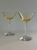 Two topaz colored glass coupe-style cocktail glasses with a clear tiered stem, shown on a light gray background .