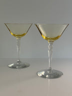 Two topaz colored glass coupe-style cocktail glasses with a clear tiered stem, shown on a light gray background .