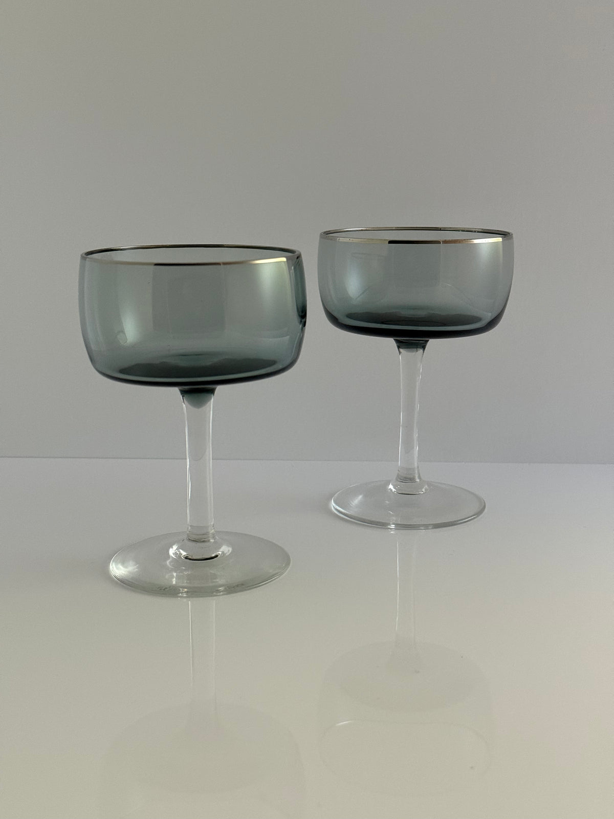 Two elegant coupe glasses with a smoky translucent gray-blue tint and a thin gold rim, displayed on a reflective white surface against a light gray background.