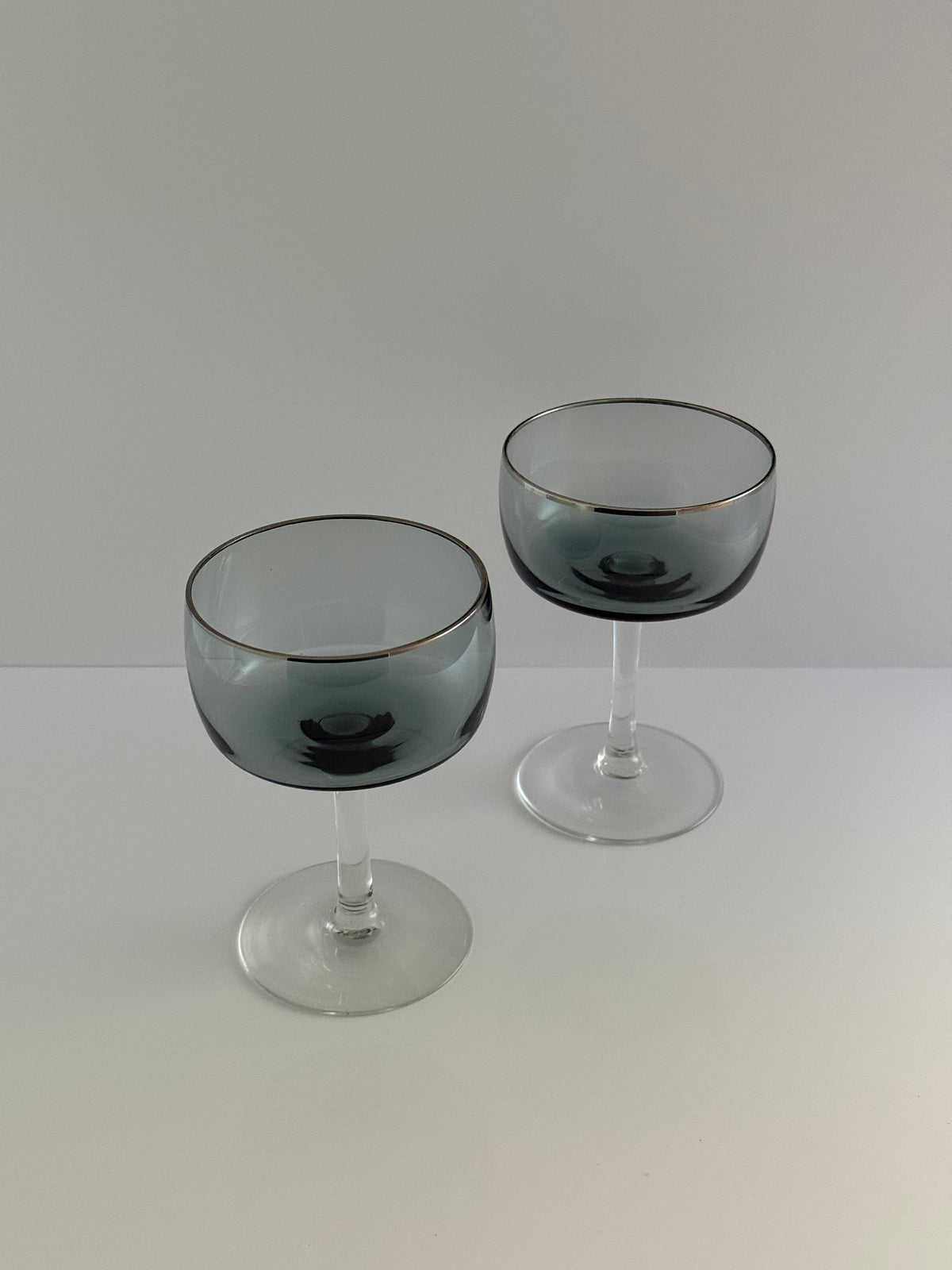 Two clear stemmed glasses with a smoky gray-blue tint and a thin gold rim, displayed on a clean white surface against a light gray background.