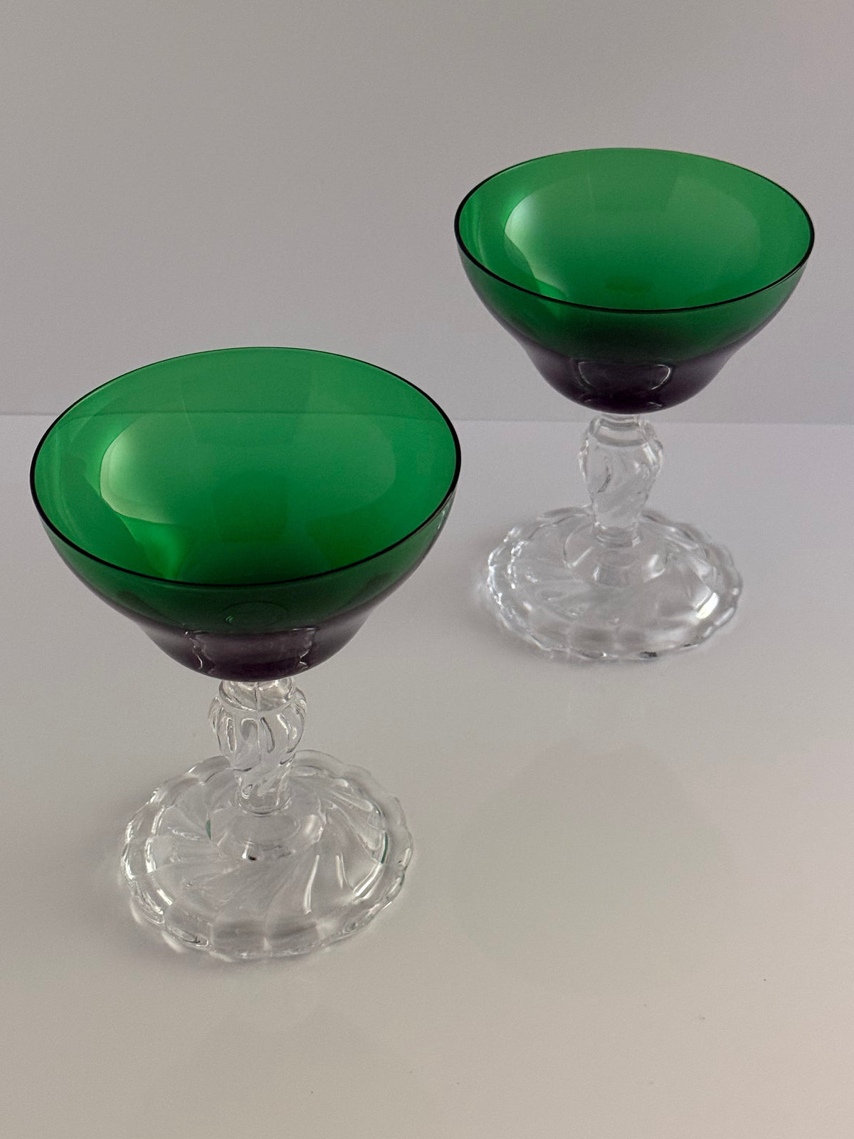 Two coupe glasses with vibrant green translucent bowls and clear faceted crystal stems and bases, set against a neutral light gray background.