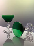 A pair of elegant coupe-style glasses with deep emerald green bowls and clear faceted crystal stems and bases, displayed on a reflective surface against a soft gradient background.