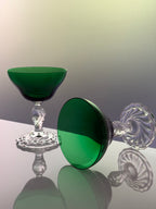 A pair of elegant coupe-style glasses with deep emerald green bowls and clear faceted crystal stems and bases, displayed on a reflective surface against a soft gradient background.