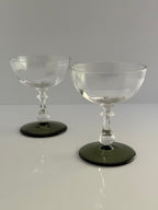 Two clear stemmed coupe glasses with dark green circular bases, photographed on a light gray studio background.