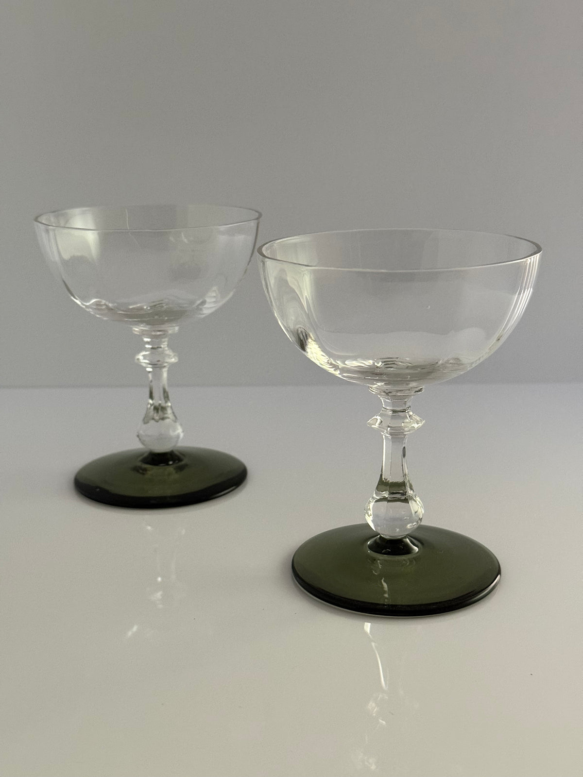 Two clear stemmed coupe glasses with dark green circular bases, photographed on a light gray studio background.