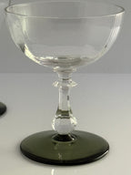 A clear glass coupe-style goblet with a dark green tinted circular base, photographed on a light gray studio background.