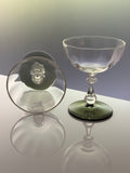 A clear glass coupe glass with a dark green tinted base, shown standing upright alongside its matching clear glass lid on a reflective white surface.