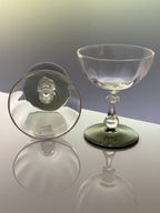 A clear glass coupe glass with a dark green tinted base, shown standing upright alongside its matching clear glass lid on a reflective white surface.