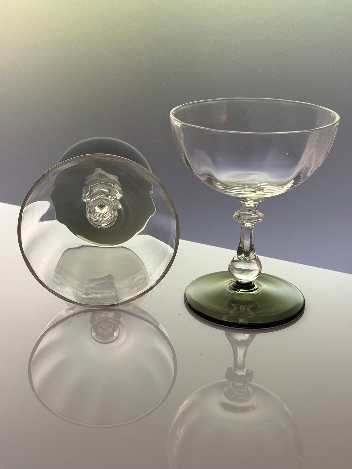 A clear glass coupe glass with a dark green tinted base, shown standing upright alongside its matching clear glass lid on a reflective white surface.