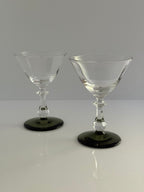 Two clear stemmed coupe glasses with dark green circular bases, displayed on a white reflective surface against a light gray background.