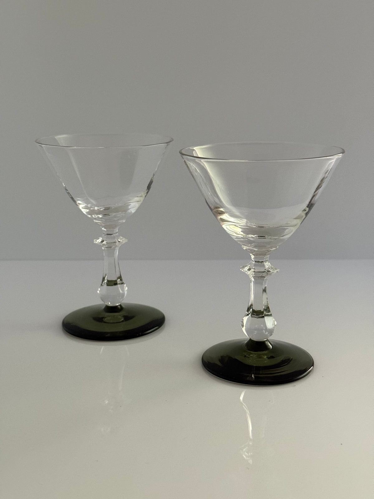 Two clear stemmed coupe glasses with dark green circular bases, displayed on a white reflective surface against a light gray background.