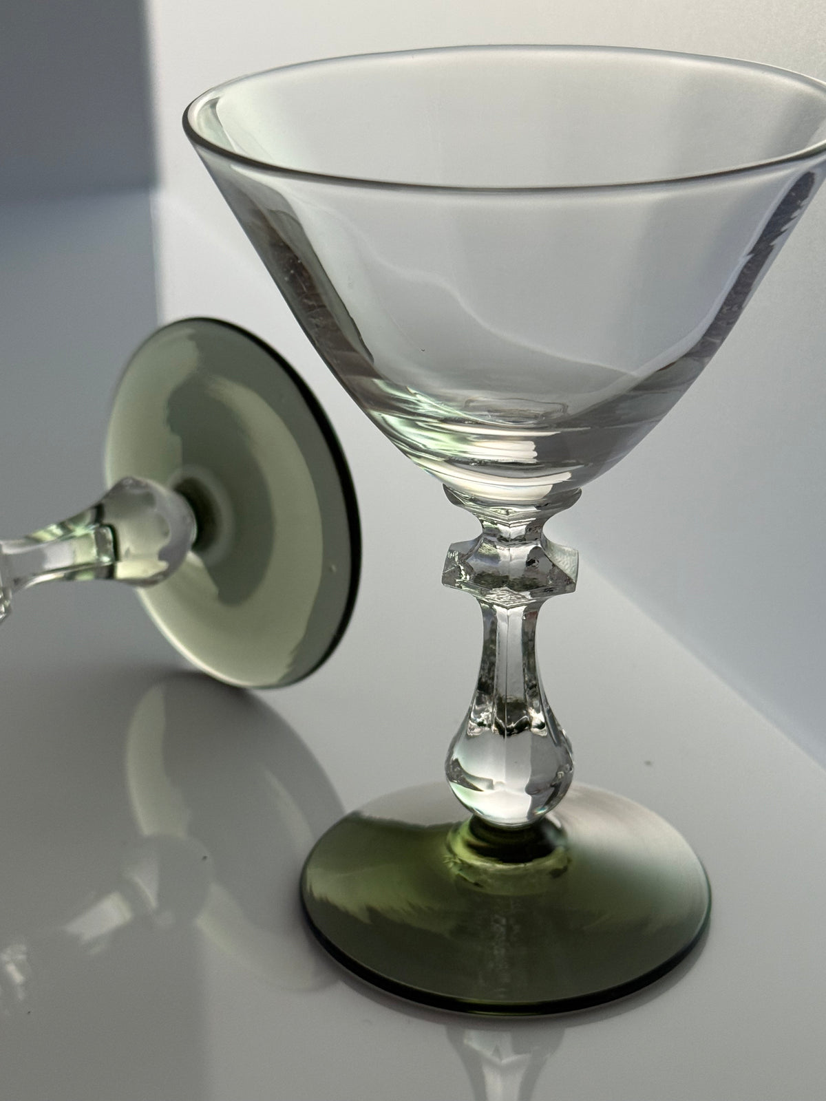 A clear glass cocktail martini glass with a green-tinted circular base and matching green-tinted stem, shown on a reflective white surface with its base removed and placed behind it.